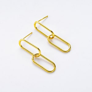 Puede incluir: Un par de pendientes de color dorado con un diseño ovalado enlazado. Cada pendiente presenta dos óvalos conectados.