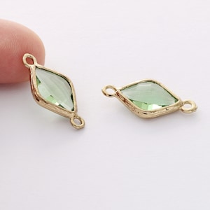 Könnte beinhalten: Zwei goldfarbene Verbindungsanhänger mit einer grünen, facettierten Glasdiamantform in der Mitte. Die Anhänger haben an beiden Seiten zwei Schlaufen zum Anbringen an Schmuck.