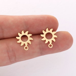 May include: Two gold-toned metal sun charm pendants with a loop at the bottom. Each charm features a sun design with a circle in the center and rays extending outward.