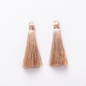 Pode incluir: Dois pingentes bege com detalhes dourados. Os pingentes são feitos de um material macio e sedoso e têm um visual delicado e feminino.