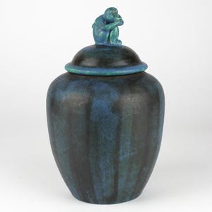 Könnte beinhalten: Eine blaue und schwarze Keramikvase mit einem Deckel, auf dem sich eine kleine blaue Affenfigur befindet. Die Vase hat eine strukturierte, streifige Glasur.