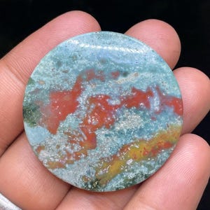 Puede incluir: Una piedra de &aacute;gata redonda, pulida, de color verde, rojo y dorado con un patr&oacute;n de remolino.