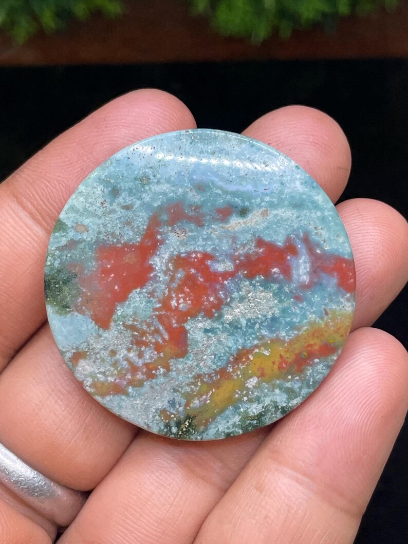 Puede incluir: Una piedra de &aacute;gata redonda, pulida, de color verde, rojo y dorado con un patr&oacute;n de remolino.