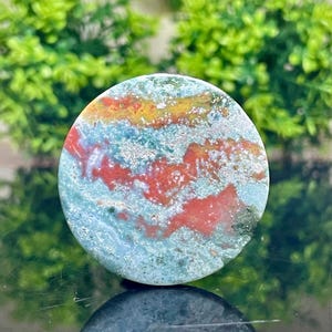 Puede incluir: Una piedra de jaspe redonda, pulida, verde, roja y dorada con un patr&oacute;n de remolino. La piedra tiene un di&aacute;metro de aproximadamente 5 cm.