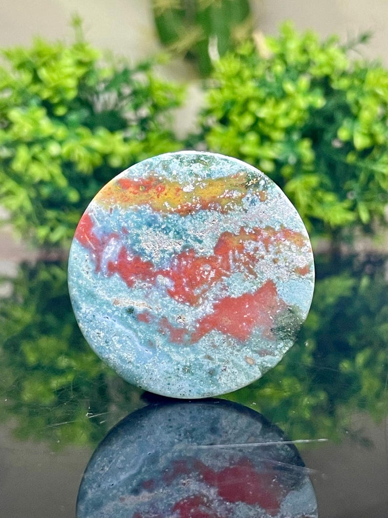 Puede incluir: Una piedra de jaspe redonda, pulida, verde, roja y dorada con un patr&oacute;n de remolino. La piedra tiene un di&aacute;metro de aproximadamente 5 cm.