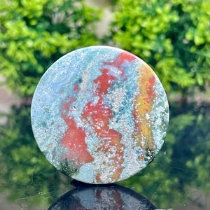 Puede incluir: Una piedra de &aacute;gata redonda, pulida, verde, roja y amarilla con un patr&oacute;n de remolino. La piedra tiene un di&aacute;metro de aproximadamente 5 cm.