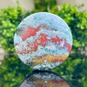 Puede incluir: Una piedra redonda y pulida con un patr&oacute;n de remolino de verde, azul, rojo y dorado. La piedra tiene un aspecto natural y org&aacute;nico.