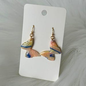 Puede incluir: Un par de pendientes con forma de mariposa con ganchos dorados. Los pendientes están hechos de un material veteado en tonos rosa, amarillo, azul y blanco. Los pendientes se muestran en una tarjeta blanca.