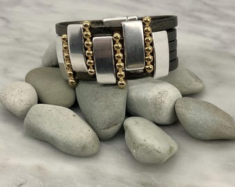 May include: A black leather bracelet with silver and gold accents. The bracelet has a geometric design with rectangular silver pieces and gold beads.