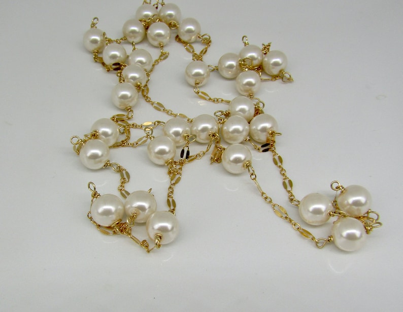 May include: A long, elegant necklace featuring large, creamy white faux pearls strung on delicate gold-colored chains. The pearls are evenly spaced, creating a classic and timeless design. The necklace is displayed against a plain white background.
