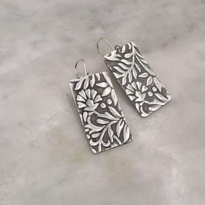May include: A pair of rectangular silver earrings featuring a raised floral and leaf pattern against a dark, oxidized background. Each earring is suspended from a simple silver ear wire, showcasing a detailed botanical relief design on the metal surface.