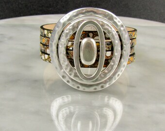May include: A silver bracelet with a hammered metal circle design. The bracelet is made of multiple layers of leather with a black and white patterned design with hints of gold.