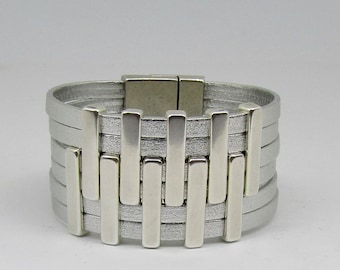 May include: A silver leather bracelet with a magnetic clasp and a silver metal design. The bracelet is made of multiple layers of leather and has a modern, geometric look.