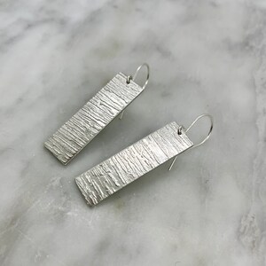 May include: A pair of silver dangle earrings with a textured rectangular design.