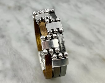 May include: A multi-layered leather bracelet with silver and gold accents. The bracelet features a repeating pattern of small silver beads and rectangular silver plates.