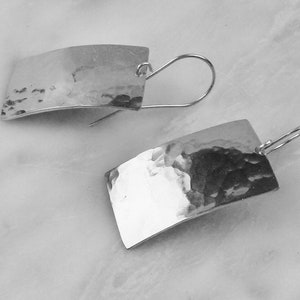 May include: A pair of silver rectangular earrings with a hammered texture. The earrings have a curved shape and are suspended from a simple hook. The earrings are displayed on a white marble surface.
