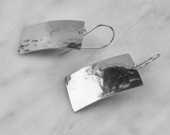 May include: A pair of silver rectangular earrings with a hammered texture. The earrings have a curved shape and are suspended from a simple hook. The earrings are displayed on a white marble surface.