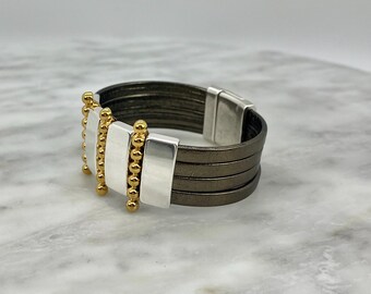 May include: A multi-layered brown leather bracelet with silver and gold accents. The bracelet features a row of gold beads and silver rectangular pieces.