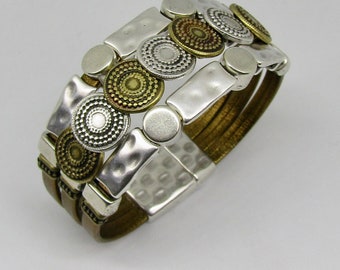 May include: A multi-layered brown leather bracelet with silver and gold metal accents. The bracelet features a series of round, decorative metal pieces with a hammered texture.