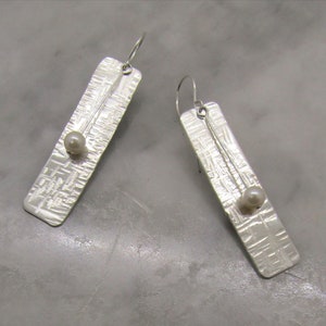 May include: A pair of silver dangle earrings with a textured rectangular design and a single white pearl on each earring.