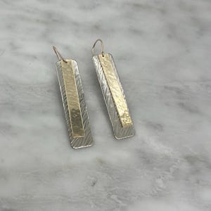 May include: A pair of long, rectangular earrings with a hammered silver finish and a gold bar accent. The earrings have a modern, minimalist design.
