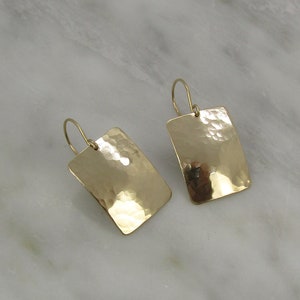 May include: A pair of gold-colored rectangular earrings with a hammered texture. The earrings have a simple hook design and reflect light, creating a shiny appearance. The earrings are displayed on a marble surface.