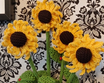 Puede incluir: Un ramo artesanal de girasoles de ganchillo en un jarrón blanco. Los girasoles tienen pétalos amarillos y centros marrones, con tallos y hojas verdes. Pequeñas mariquitas rojas están unidas a las hojas. El jarrón tiene una superficie texturizada.