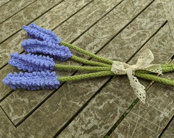 Puede incluir: Un conjunto de tres tallos de lavanda de ganchillo con cabezas de flores moradas texturizadas y tallos largos verdes. Los tallos están agrupados y atados con una cinta de encaje color crema con un delicado patrón calado.