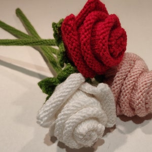 Könnte beinhalten: Drei gestrickte Rosen in Rot, Weiß und Rosa, mit grünen Stielen und Blättern. Die handgefertigten Blumen sind zusammen angeordnet und zeigen die detaillierte Textur des Garns und die realistische Rosenform.