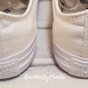 May include: A pair of white canvas Converse All-Star sneakers with a pink inscription on the side that reads "27.05.21".
