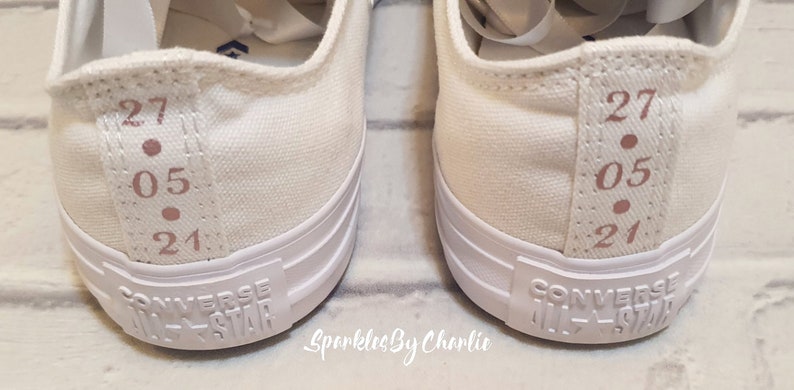 May include: A pair of white canvas Converse All-Star sneakers with a pink inscription on the side that reads "27.05.21".