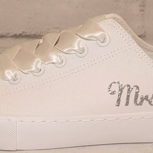 May include: White canvas sneakers with a satin ribbon tied around the laces. The shoes have a sparkly embellishment on the toe and are personalized with the text "Mrs. G" in silver glitter.