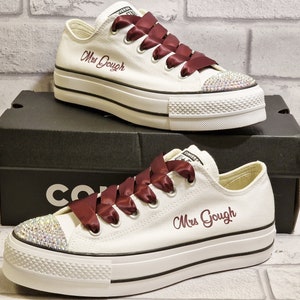 Puede incluir: Zapatillas blancas de lona de caña alta con cordones de satén granate y un nombre bordado en burdeos "Mrs. Gough" en el lateral. Las zapatillas tienen un adorno de pedrería en la puntera.