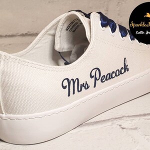 May include: White canvas low-top sneakers with blue satin laces and the text "Mrs Peacock" embroidered in navy blue.