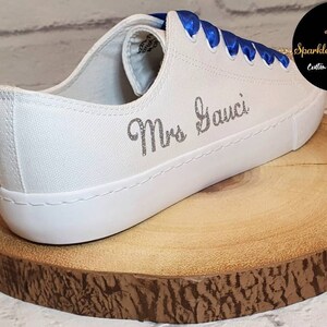 May include: White canvas sneakers with blue satin laces and the text "Mrs. Gauci" in silver glitter on the side.