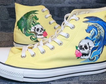 May include: A pair of yellow high-top sneakers with a dragon and skull design. One shoe features a green dragon and the other a blue dragon. Both shoes have a skull with a pink rose.