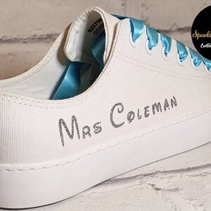May include: White canvas sneakers with a blue satin ribbon tied around the laces. The shoes have a silver glitter inscription that reads "Mrs. Coleman".