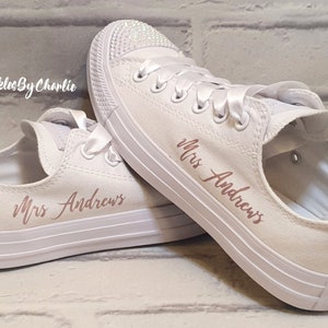 May include: White canvas sneakers with rose gold lettering that says "Mrs. Andrews" on both shoes. The shoes have white satin laces and a white brick wall background.