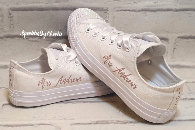 May include: White canvas sneakers with rose gold lettering that says "Mrs. Andrews" on both shoes. The shoes have white satin laces and a white brick wall background.
