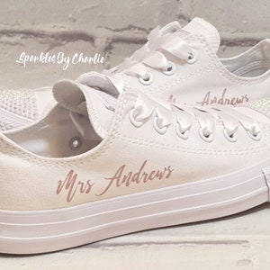 May include: White canvas sneakers with a rose gold inscription that reads "Mrs. Andrews". The sneakers have white satin laces and a white rubber sole. The sneakers are decorated with rhinestones.