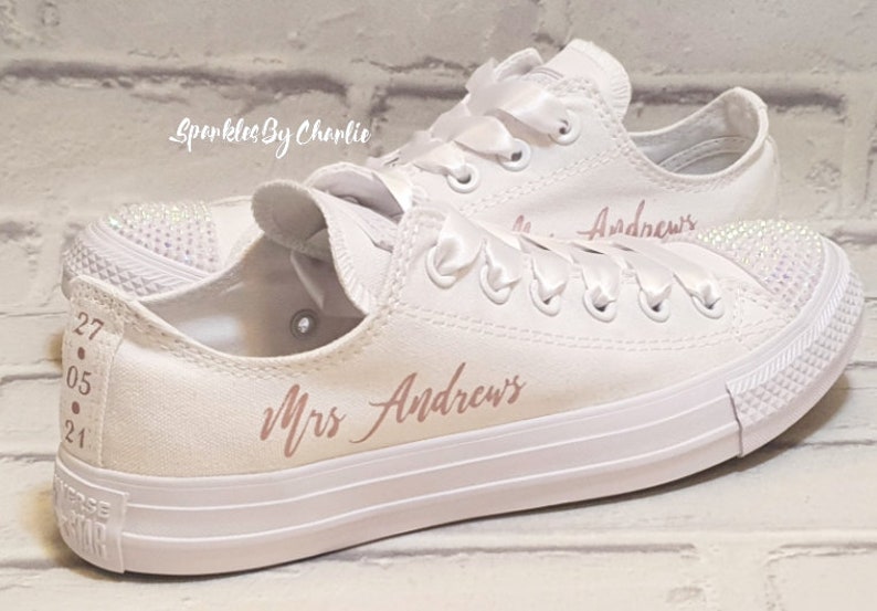 May include: White canvas sneakers with a rose gold inscription that reads "Mrs. Andrews". The sneakers have white satin laces and a white rubber sole. The sneakers are decorated with rhinestones.
