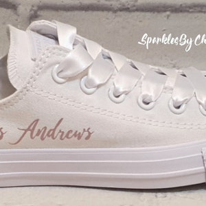 May include: White canvas low-top sneakers with a satin ribbon tied around the laces. The shoes are personalized with the text "Mrs. Andrews" in rose gold lettering and have a crystal embellishment on the toe.