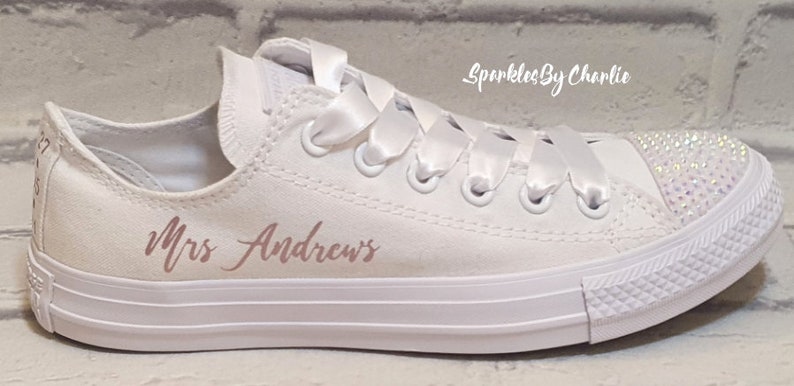May include: White canvas low-top sneakers with a satin ribbon tied around the laces. The shoes are personalized with the text "Mrs. Andrews" in rose gold lettering and have a crystal embellishment on the toe.