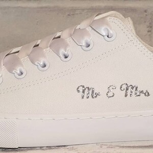 May include: White canvas sneakers with a blue rhinestone embellishment on the toe. The shoes are personalized with the text "Mr. & Mrs. Twigge" in silver glitter.