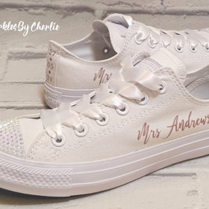 May include: White canvas sneakers with a rose gold inscription that reads "Mrs. Andrews". The sneakers have white satin laces and a rhinestone embellishment on the toe.
