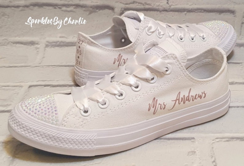 May include: White canvas sneakers with a rose gold inscription that reads "Mrs. Andrews". The sneakers have white satin laces and a rhinestone embellishment on the toe.