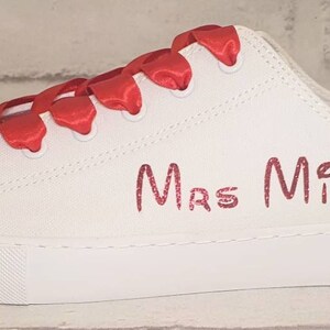 May include: White canvas sneakers with red satin laces and a sparkly rhinestone embellishment on the toe. The shoes are personalized with the text "Mrs Mills" in red.