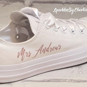 May include: White canvas low-top sneakers with a rose gold inscription that reads "Mrs. Andrews" and a row of rhinestones on the toe. The date "27.05.21" is written on the back of the shoe.