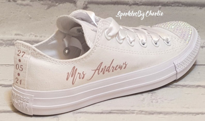 May include: White canvas low-top sneakers with a rose gold inscription that reads "Mrs. Andrews" and a row of rhinestones on the toe. The date "27.05.21" is written on the back of the shoe.