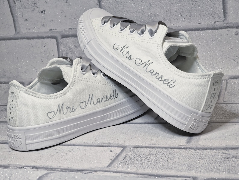 May include: A pair of white canvas sneakers with silver ribbon laces. The sneakers are personalized with the text "Mrs. Mansell" and a heart symbol.